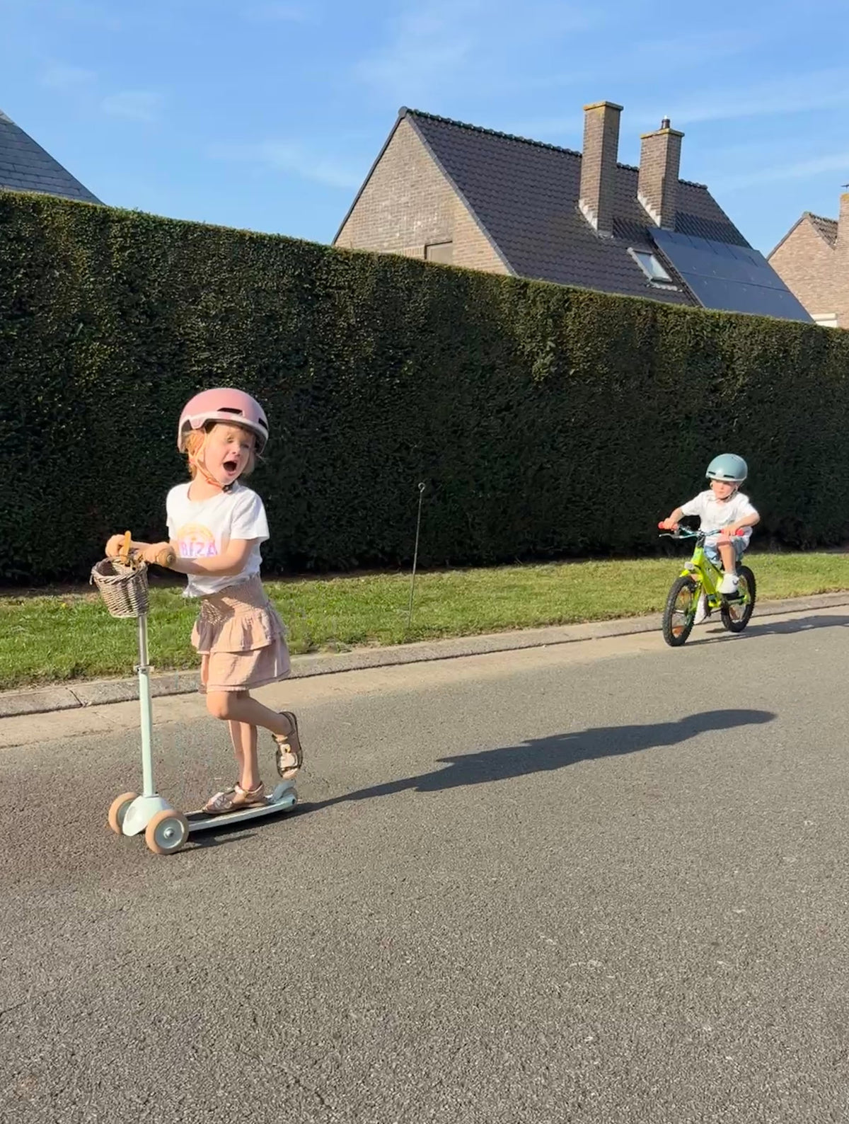 Meisje met roze helm op een step en jongen met helm op de fiets in een zonnige straat